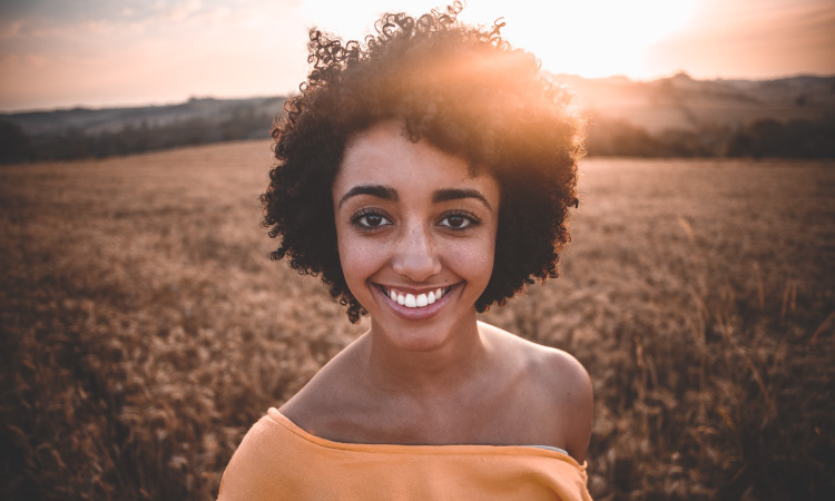 Curly-haired woman wearing a yellow blouse smiles in a wheat field after teeth whitening
