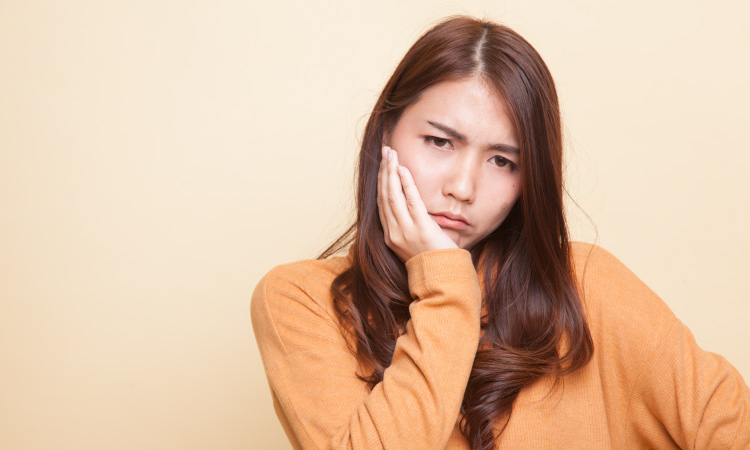 Brunette woman wearing a yellow sweater cringes and touches her jaw due to TMJ pain