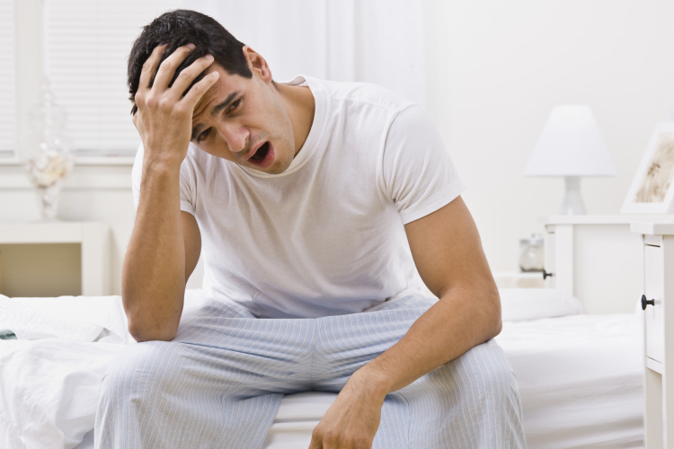 Brunette man in his pajamas sits on the edge of his bed with a headache after a night of grinding his teeth