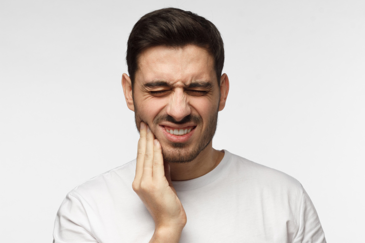 Dark-haired man cringes in pain and touches his cheek due to a toothache dental emergency