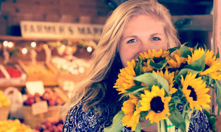 Blonde woman at a farmers market covers her mouth with a bouquet of sunflowers because she has gum disease