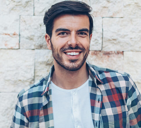 A man with a plaid shirt and veneers smiling in front of a brick wall