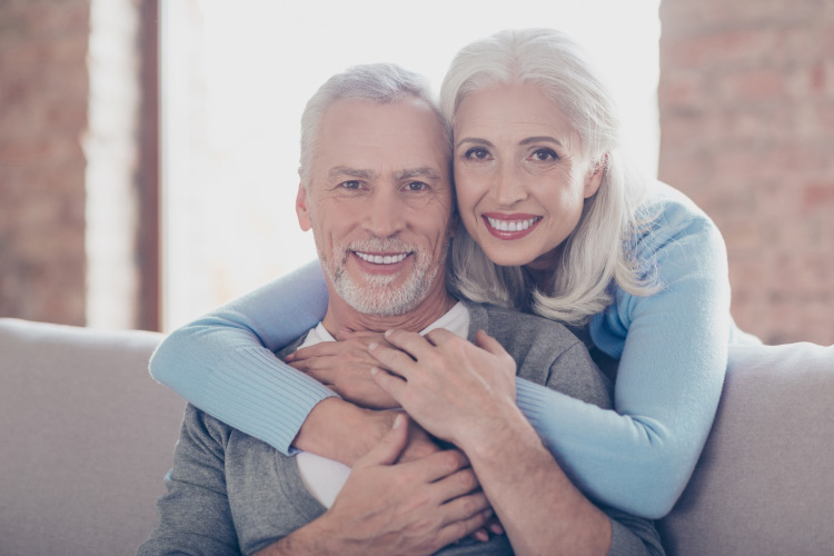 Older white-haired couple with dental implants embrace while sitting on a couch